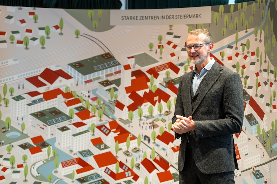 Ein Mann im Anzug steht lächelnd vor einer großen illustrierten Landkarte mit Gebäuden und Bäumen. Oben steht der Schriftzug "STARKE ZENTREN IN DER STEIERMARK" und weist auf das Autohaus Gaberszik Graz und 100 Jahre Autohaus Gaberszik hin.