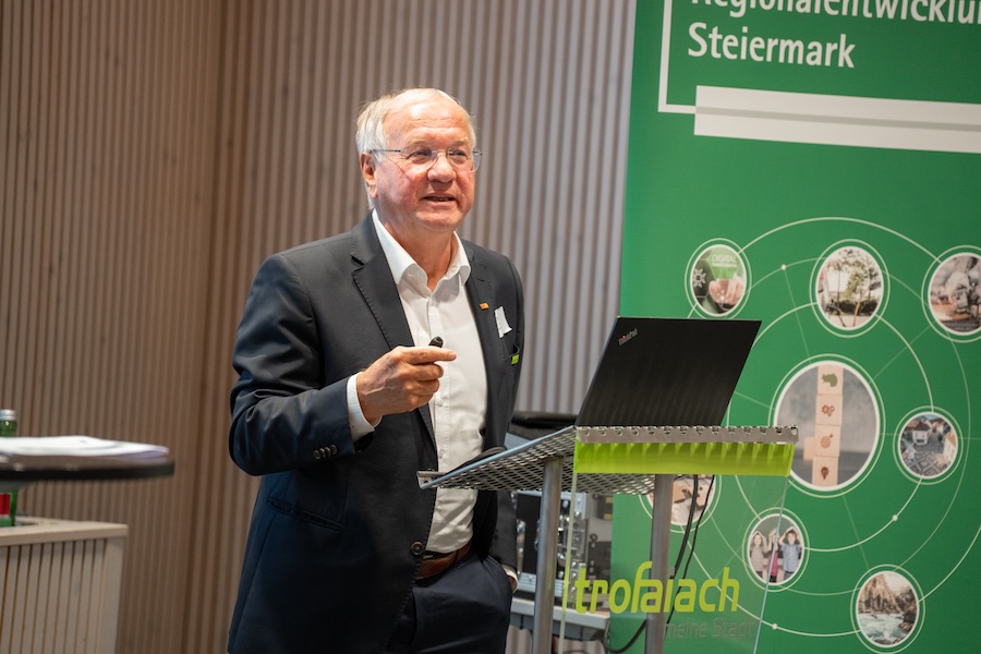 Ein älterer Mann im Anzug spricht mit einer Fernbedienung in der Hand, er steht an einem Podium mit einem Laptop. Hinter ihm ist ein grünes Banner mit kreisförmigen Bildern, der Aufschrift "Regionalentwicklung Steiermark" und dem Landeswappen Steiermark zu sehen.