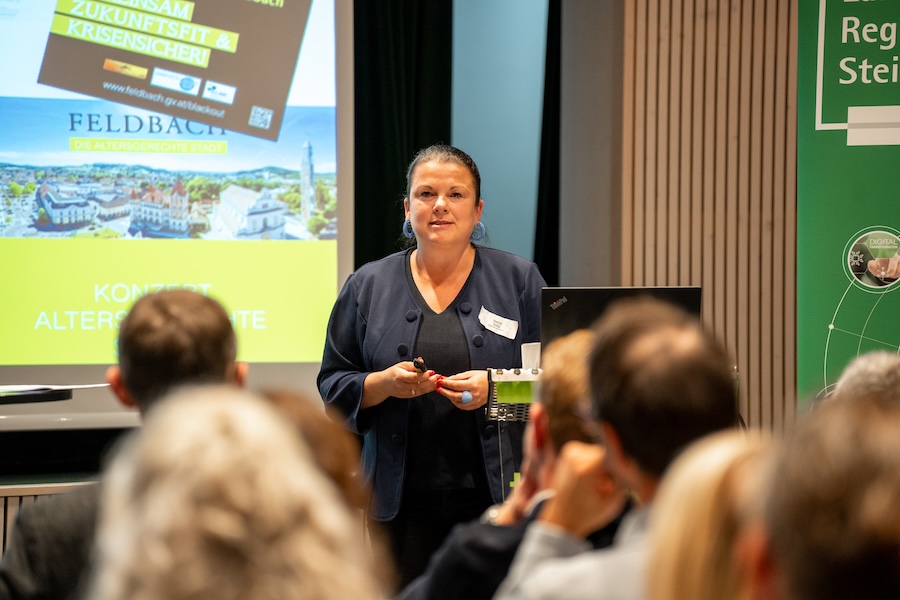 Eine Frau steht vor einem sitzenden Publikum, spricht und hält ein Mikrofon. Hinter ihr zeigt ein Bildschirm eine Präsentation mit der Überschrift "Feldbach" und "100 Jahre Autohaus Gaberszik" sowie ein grünes Werbebanner.
