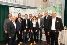 Eine Gruppe von elf Männern und Frauen steht lächelnd vor einer farbenfrohen Kulisse mit Landkarten, Bäumen und dem Landeswappen Steiermark und feiert 100 Jahre Autohaus Gaberszik. Die meisten tragen Business-Casual-Kleidung und Namensschilder.