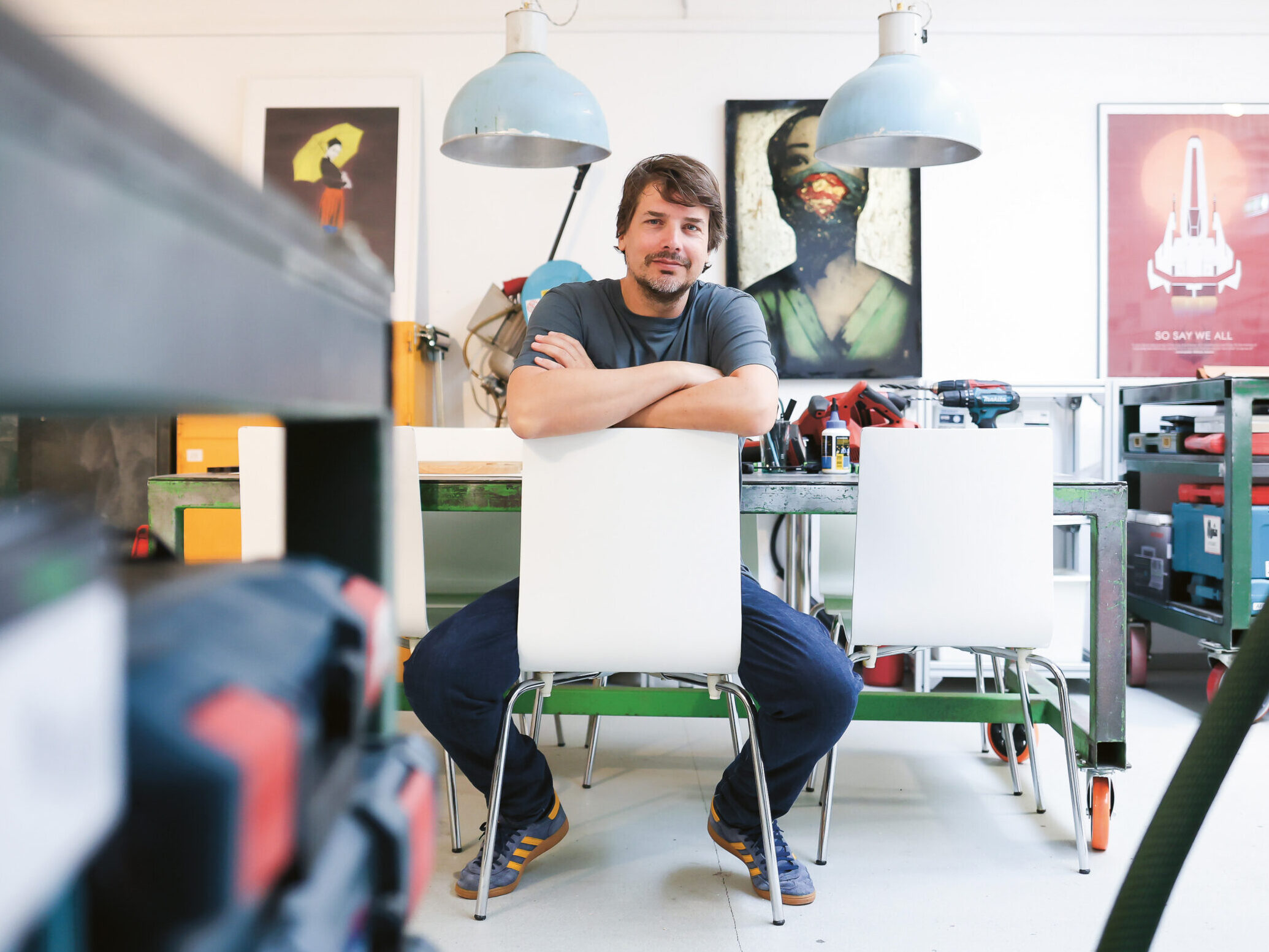 Ein Mann sitzt auf einem weißen Stuhl an einem Tisch in einer hellen, modernen Werkstatt mit Werkzeugen und Geräten um ihn herum. An der Wand dahinter hängen drei Kunstposter, und von der Decke hängen zwei Pendelleuchten.