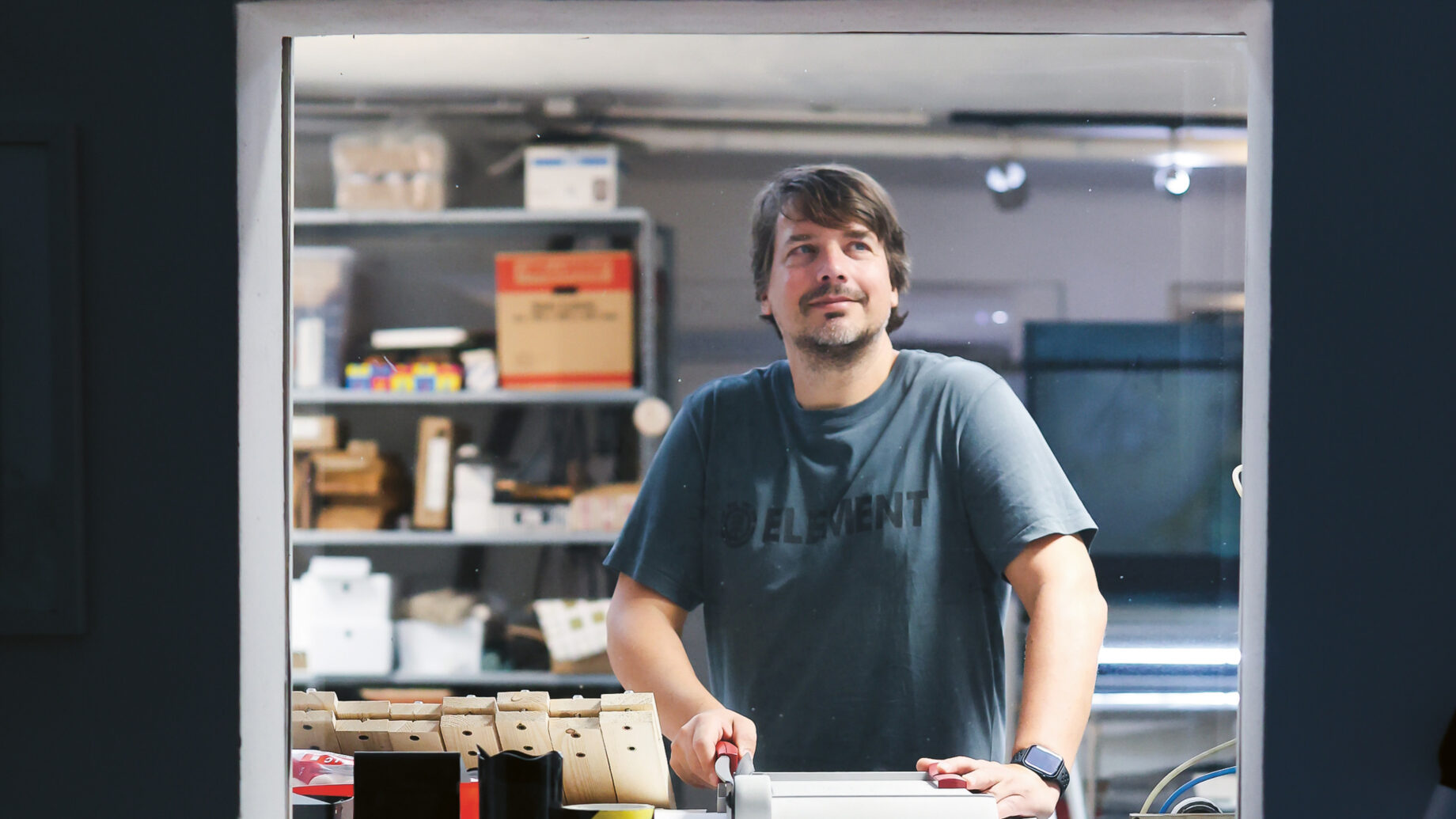 Ein Mann steht hinter einem Tresen in einem gut beleuchteten Arbeitsbereich und schaut in die Kamera. Auf dem Tresen stehen verschiedene Gegenstände, darunter ein Buch, eine kleine Figur und ein rotes anatomisches Modell. Im Hintergrund befinden sich Regale und Geräte.