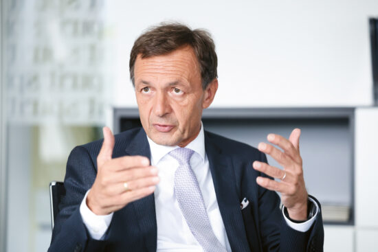 Ein Mann in einem dunklen Anzug und einer hellen Krawatte sitzt in einem Haus und spricht mit beiden Händen, während er gestikuliert. Er hat kurzes braunes Haar und einen ernsten Gesichtsausdruck. Im Hintergrund sind Regale und helle Wände zu sehen.