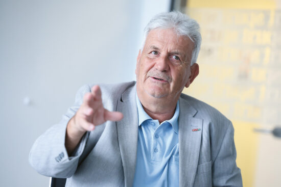 Ein älterer Mann mit grauem Haar und Schnurrbart, der einen hellgrauen Blazer und ein blassblaues Hemd trägt, gestikuliert mit seiner rechten Hand, während er in einem Haus sitzt. Der Hintergrund ist leicht unscharf, mit gelben und grauen Tönen.