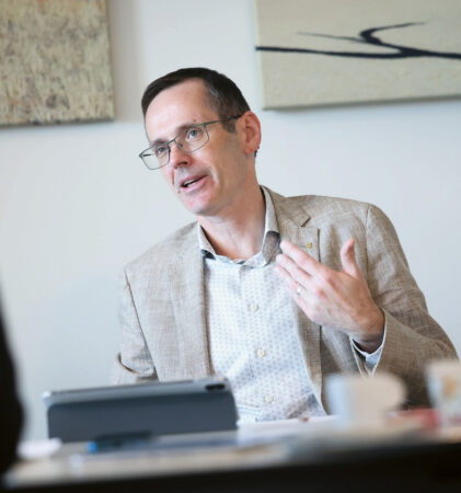 Ein Mann mit Brille und beigem Blazer sitzt an einem Schreibtisch und gestikuliert mit einer Hand, während er spricht. Auf dem Schreibtisch liegen Papiere und ein Tablet, und an der Wand hinter ihm hängt abstrakte Kunst.