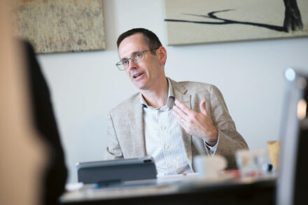 Ein Mann mit Brille und beigem Blazer sitzt an einem Schreibtisch und gestikuliert mit einer Hand, während er spricht. Auf dem Schreibtisch liegen Papiere und ein Tablet, und an der Wand hinter ihm hängt abstrakte Kunst.