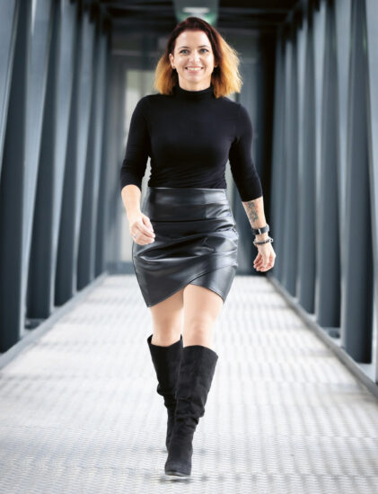 Eine Frau mit schulterlangem braunem Haar geht selbstbewusst auf einer Metallbrücke, trägt einen schwarzen Rollkragenpullover, einen schwarzen Lederrock und schwarze kniehohe Stiefel. Im Hintergrund sind geometrische Metallträger zu sehen.