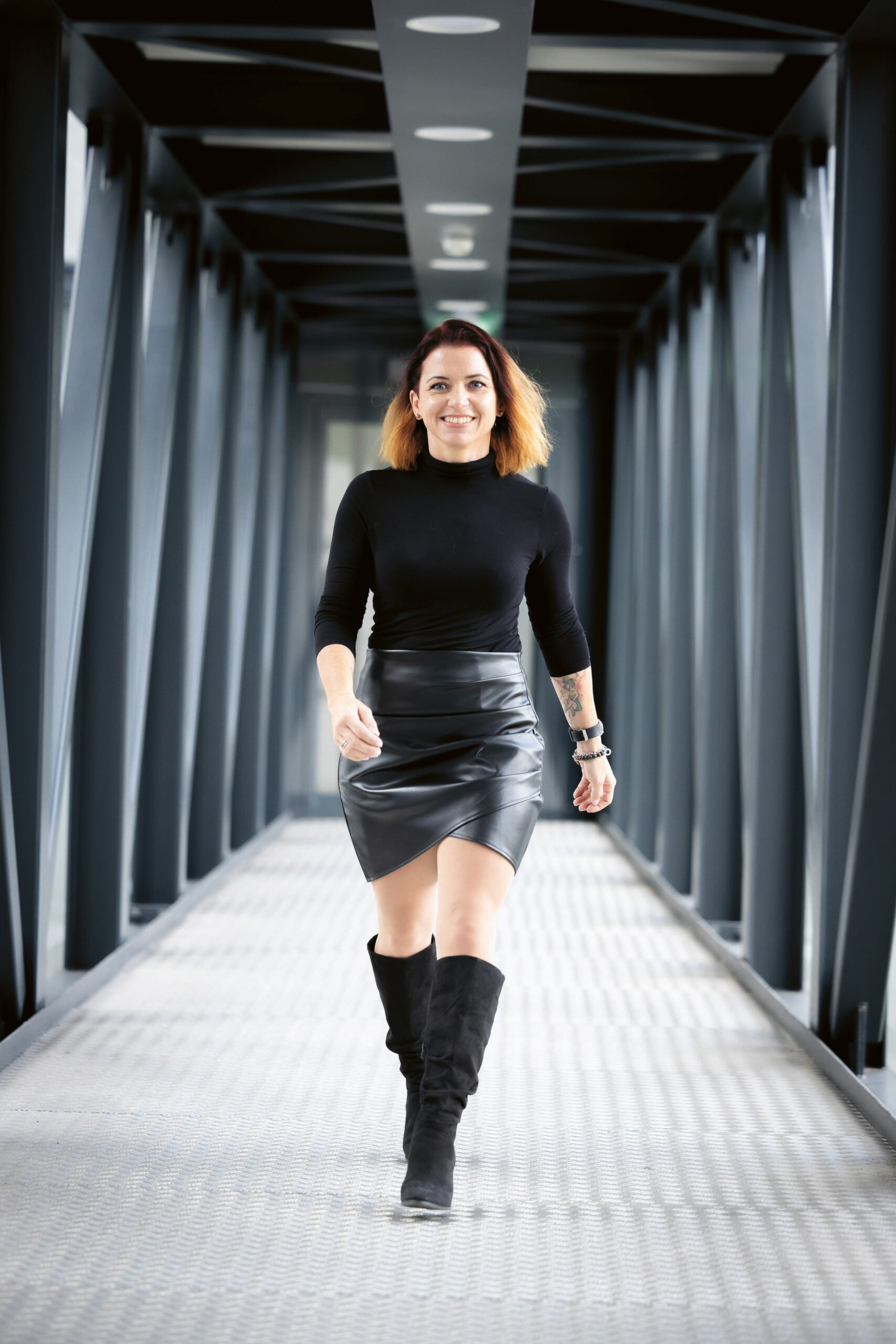 Eine Frau mit schulterlangem Haar, schwarzem Rollkragenpullover, schwarzem Lederrock und schwarzen kniehohen Stiefeln geht selbstbewusst über eine moderne Fußgängerbrücke aus Metall mit geometrischen Balken.
