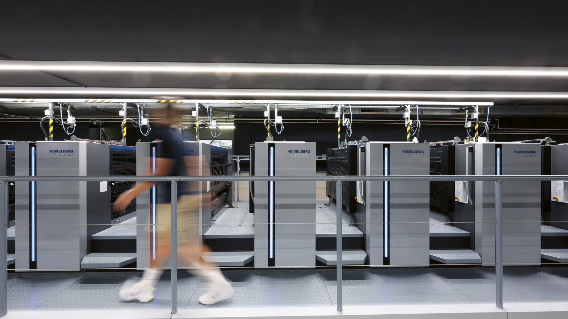 Eine verschwommene Person geht an einer Reihe großer, moderner Industriemaschinen in einem gut beleuchteten Gebäude vorbei, möglicherweise ein Rechenzentrum oder eine Druckerei, in dem Oberlichter und Metallgeländer sichtbar sind.