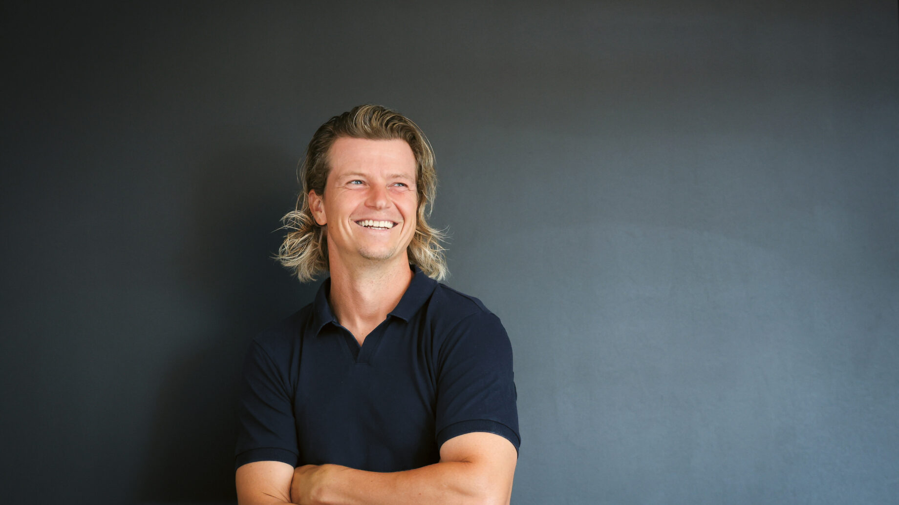 Ein lächelnder Mann mit schulterlangem blondem Haar und einem marineblauen Polohemd steht mit verschränkten Armen vor einem schlichten dunkelgrauen Hintergrund.