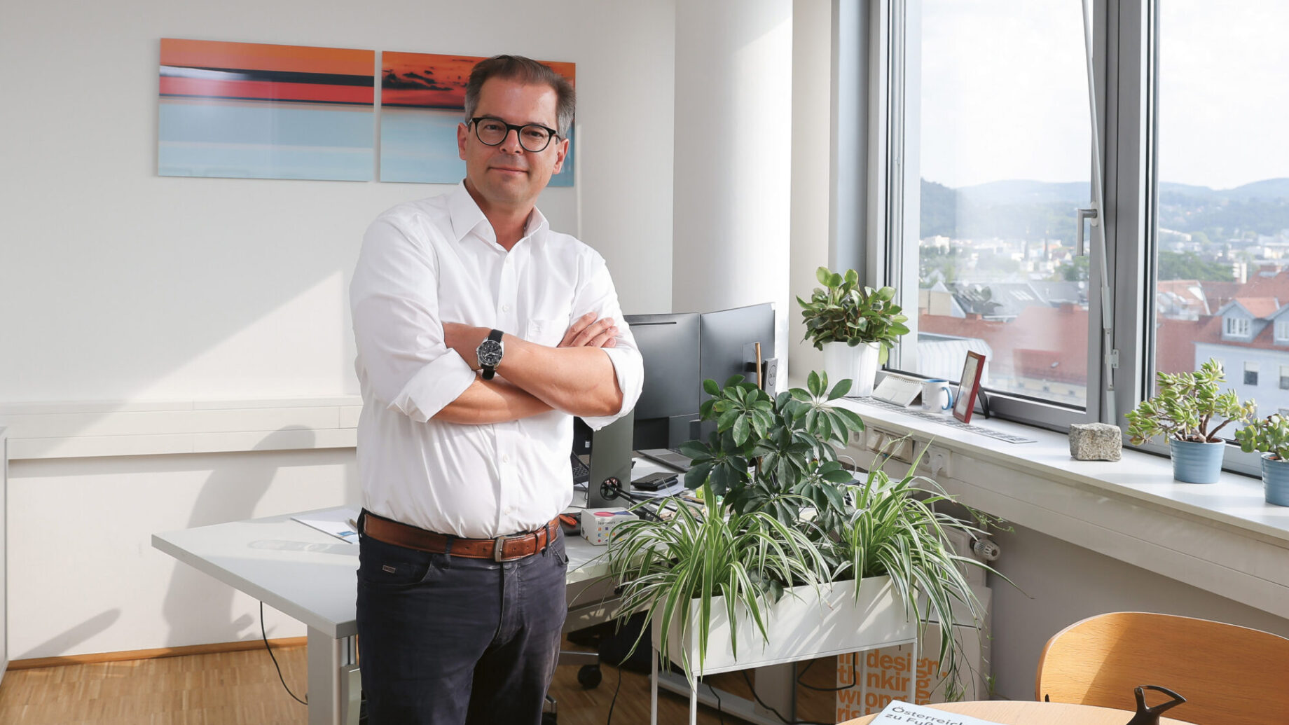 Ein Mann mit Brille und weißem Hemd steht mit verschränkten Armen in einem hellen, mit Pflanzen gefüllten Büro. Ein großes Fenster gibt den Blick auf eine Stadtlandschaft frei, und an der Wand stehen Schreibtische, Computer und Kunstwerke.