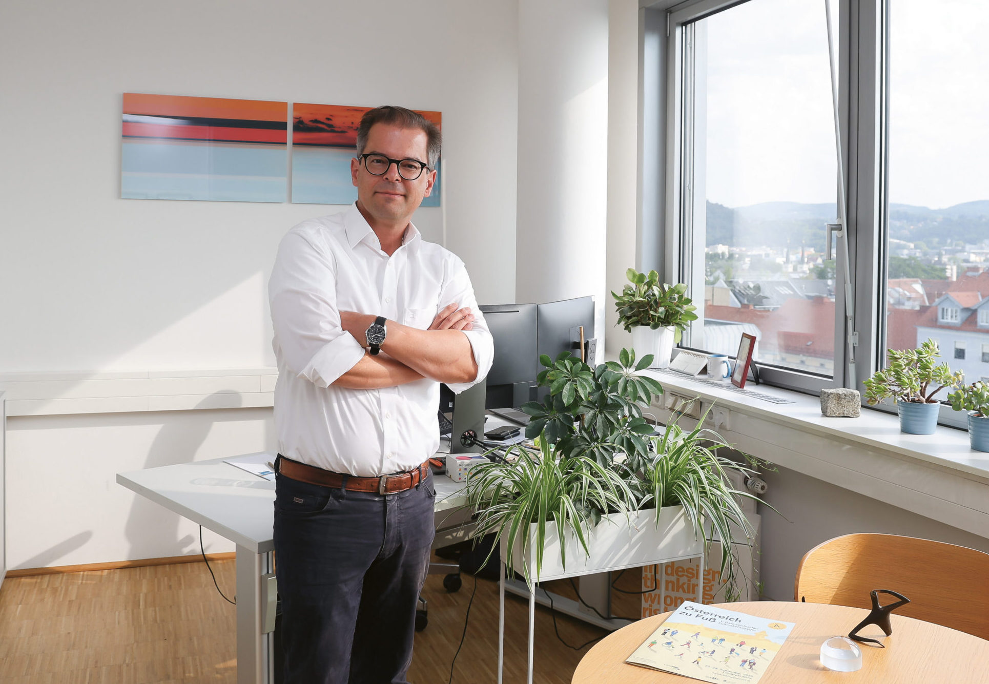 Ein Mann mit Brille und weißem Hemd steht mit verschränkten Armen in einem hellen, mit Pflanzen gefüllten Büro. Ein großes Fenster gibt den Blick auf eine Stadtlandschaft frei, und an der Wand stehen Schreibtische, Computer und Kunstwerke.