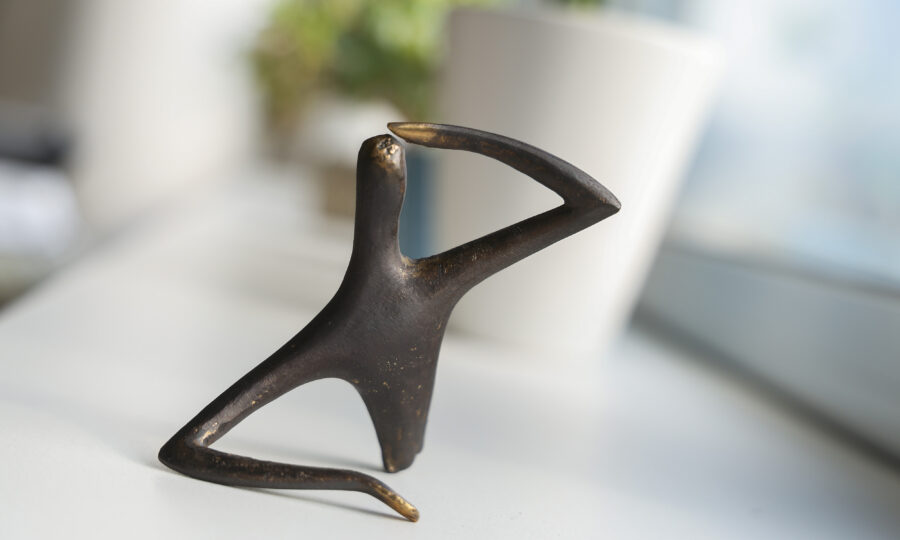 Kleine abstrakte Bronzeskulptur einer sitzenden menschlichen Figur, die einen Arm auf die Stirn gelegt hat. Sie steht auf einer weißen Fläche mit einem verschwommenen weißen Topf und einer grünen Pflanze im Hintergrund.