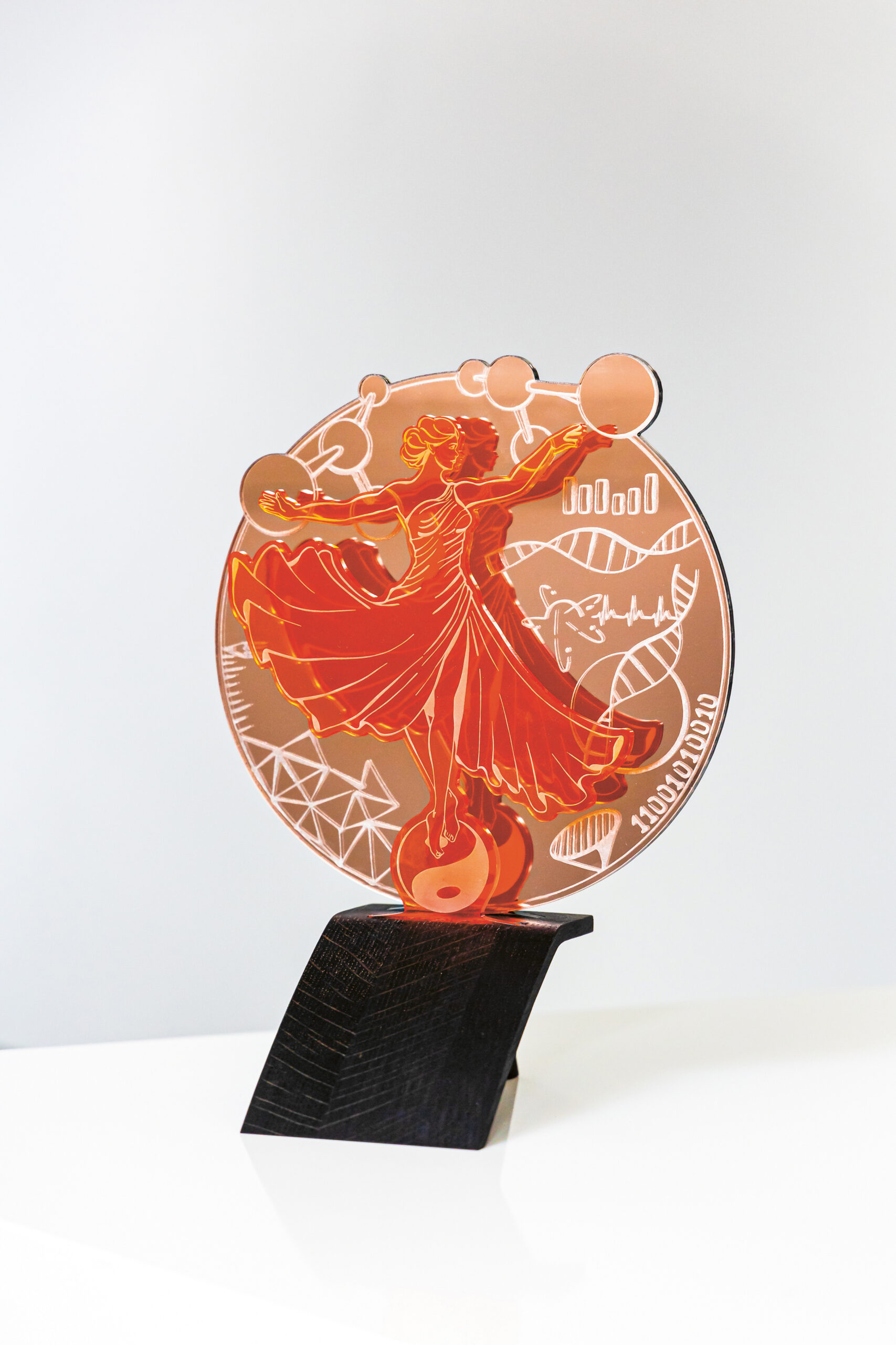 Ein orangefarbener Acryl-Award mit einer stilisierten tanzenden Frau, umgeben von wissenschaftlichen Symbolen wie DNA-Strängen, Molekülen und geometrischen Formen, montiert auf einem schwarzen Ständer.