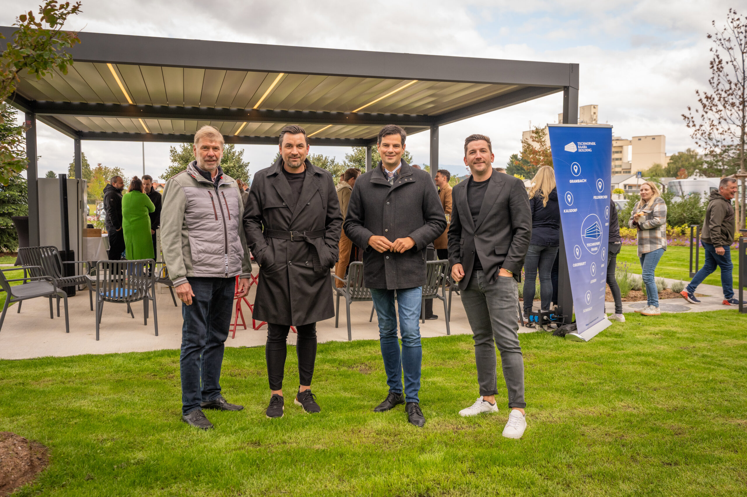 Vier Männer posieren für ein Foto auf einer Wiese vor einer modernen Pergola im Neuen Park im Technopark Raaba, mit einer Gruppe von Menschen und einem blauen Werbebanner im Hintergrund an einem bewölkten Tag.