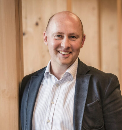 Ein Mann mit Glatze, der ein hellgestreiftes Hemd und einen strukturierten Blazer trägt, lehnt an einer Holzwand und lächelt warm in einem sanft beleuchteten, modernen Innenraum.