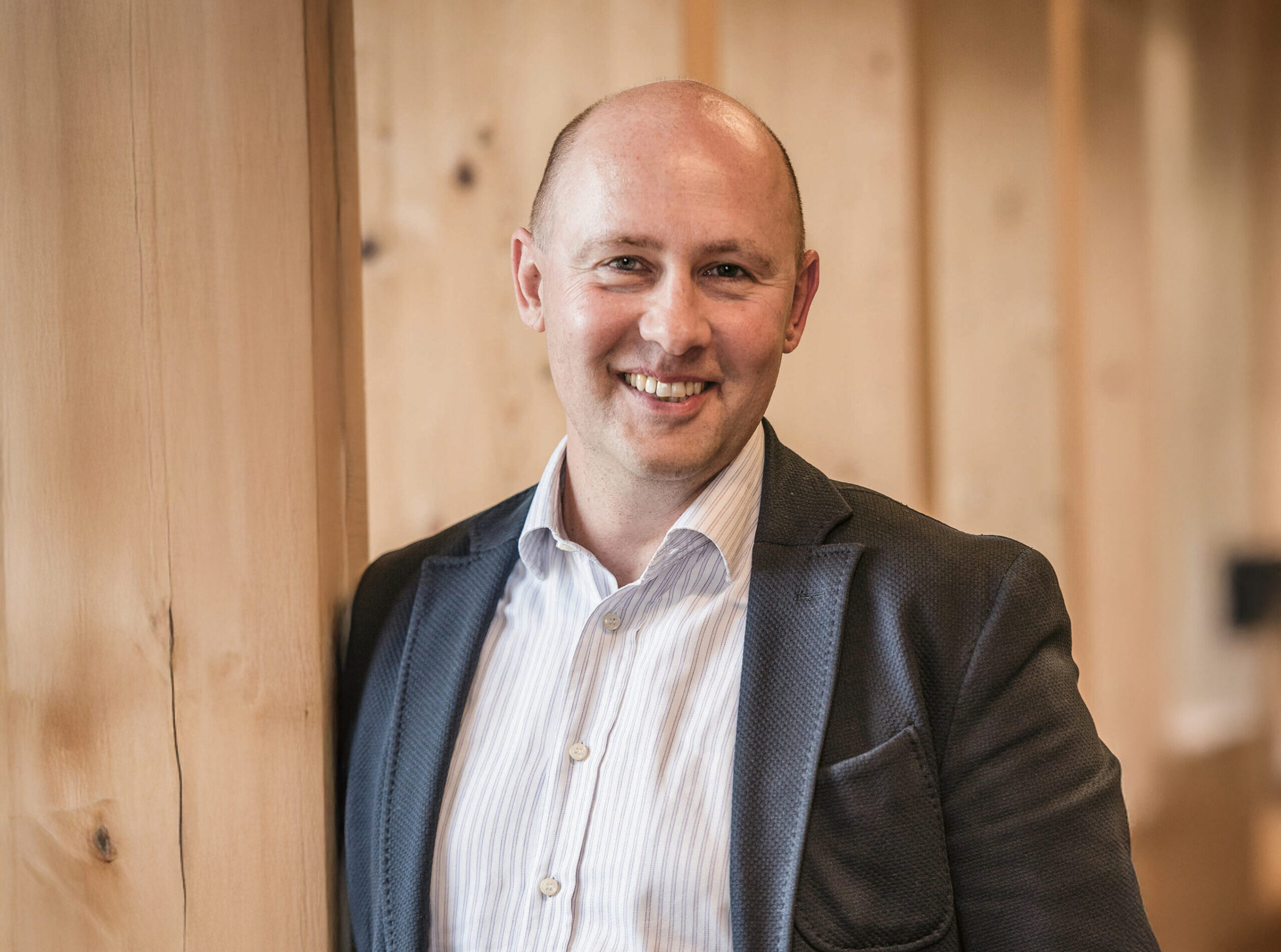Ein Mann mit Glatze, der ein hellgestreiftes Hemd und einen strukturierten Blazer trägt, lehnt an einer Holzwand und lächelt warm in einem sanft beleuchteten, modernen Innenraum.