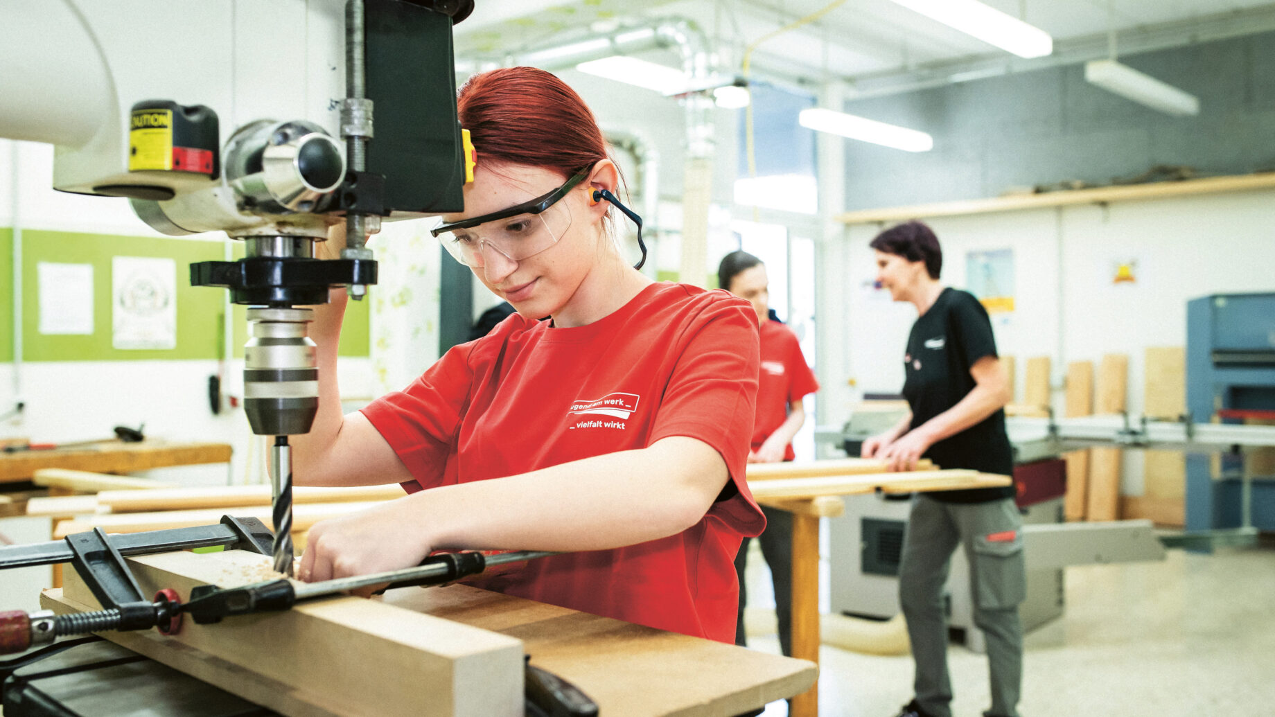 Eine Person mit Schutzbrille und rotem Hemd bedient in einer Werkstatt eine Bohrmaschine an einem Stück Holz, während zwei weitere Personen an Bänken im Hintergrund arbeiten.