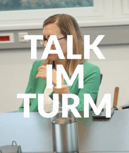 Eine Frau mit langen blonden Haaren und Brille sitzt an einem Konferenztisch und gestikuliert, während sie spricht. In großer weißer Schrift steht "TALK IM TURM". Eine Wasserflasche und Gläser stehen auf dem Tisch.