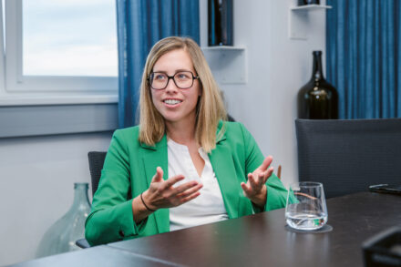 Eine Frau mit blondem Haar und Brille, die einen grünen Blazer und ein weißes Oberteil trägt, sitzt an einem Tisch und gestikuliert mit ihren Händen. Auf dem Tisch stehen ein Glas Wasser und ein Smartphone, im Hintergrund sind Regale und blaue Vorhänge zu sehen.