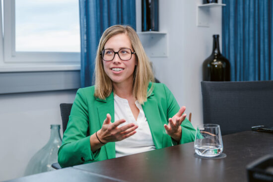 Eine Frau mit blondem Haar und Brille, die einen grünen Blazer und ein weißes Oberteil trägt, sitzt an einem Tisch und gestikuliert mit ihren Händen. Auf dem Tisch stehen ein Glas Wasser und ein Smartphone, im Hintergrund sind Regale und blaue Vorhänge zu sehen.