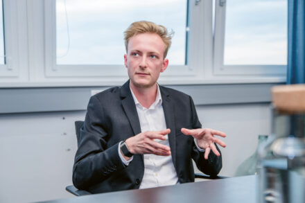 Ein Mann im Anzug sitzt an einem Schreibtisch in einem Büro und gestikuliert mit seinen Händen, während er spricht. Er hat kurzes blondes Haar und blickt leicht zur Seite. Im Hintergrund sind große Fenster zu sehen.