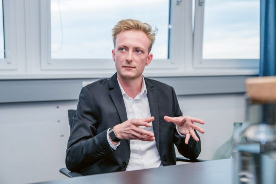 Ein Mann im Anzug sitzt an einem Schreibtisch in einem Büro und gestikuliert mit seinen Händen, während er spricht. Er hat kurzes blondes Haar und blickt leicht zur Seite. Im Hintergrund sind große Fenster zu sehen.