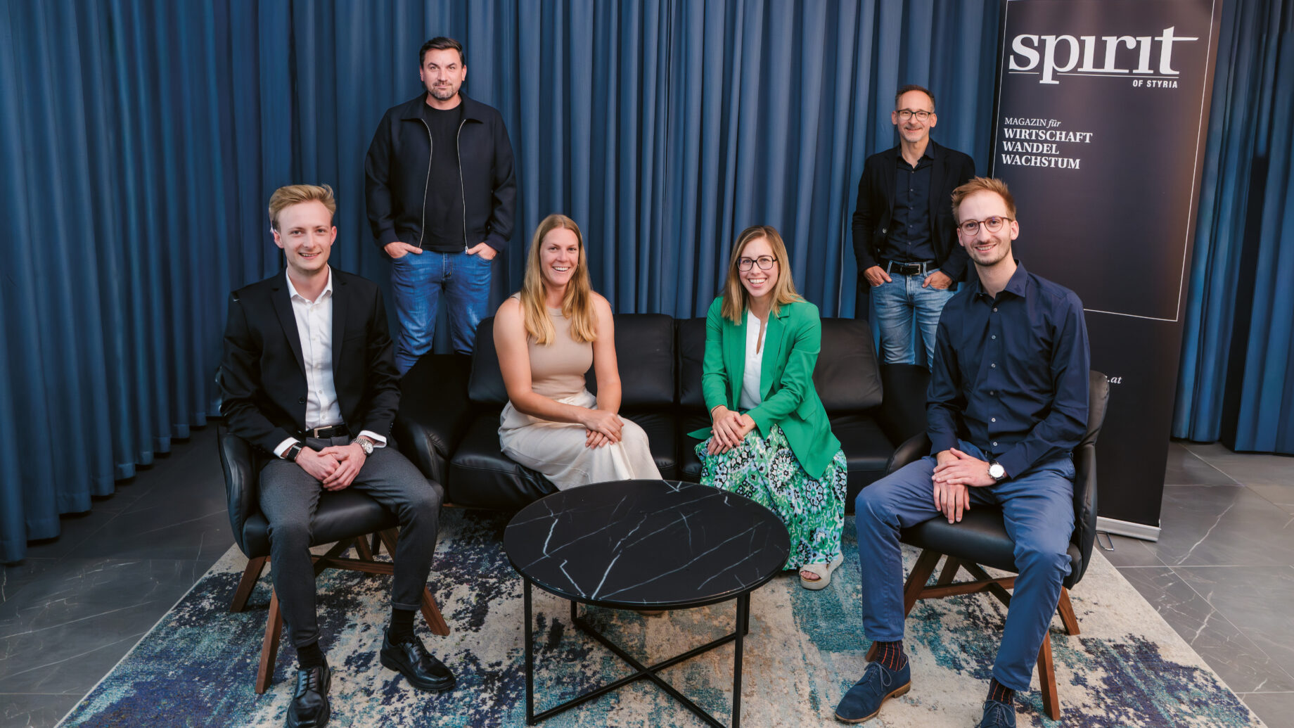 Sechs Personen, drei sitzend und drei stehend, posieren für ein Gruppenfoto in einem modernen Raum mit blauen Vorhängen, einem Teppich, einer schwarzen Couch, einem runden Marmortisch und einem "Spirit"-Banner auf der rechten Seite.