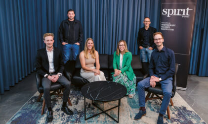 Sechs Personen, drei sitzend und drei stehend, posieren für ein Gruppenfoto in einem modernen Raum mit blauen Vorhängen, einem Teppich, einer schwarzen Couch, einem runden Marmortisch und einem "Spirit"-Banner auf der rechten Seite.