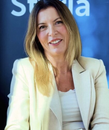 Eine Frau mit glattem, schulterlangem blondem Haar in einem cremefarbenen Blazer und weißem Oberteil lächelt, während sie vor einem blauen Hintergrund sitzt.