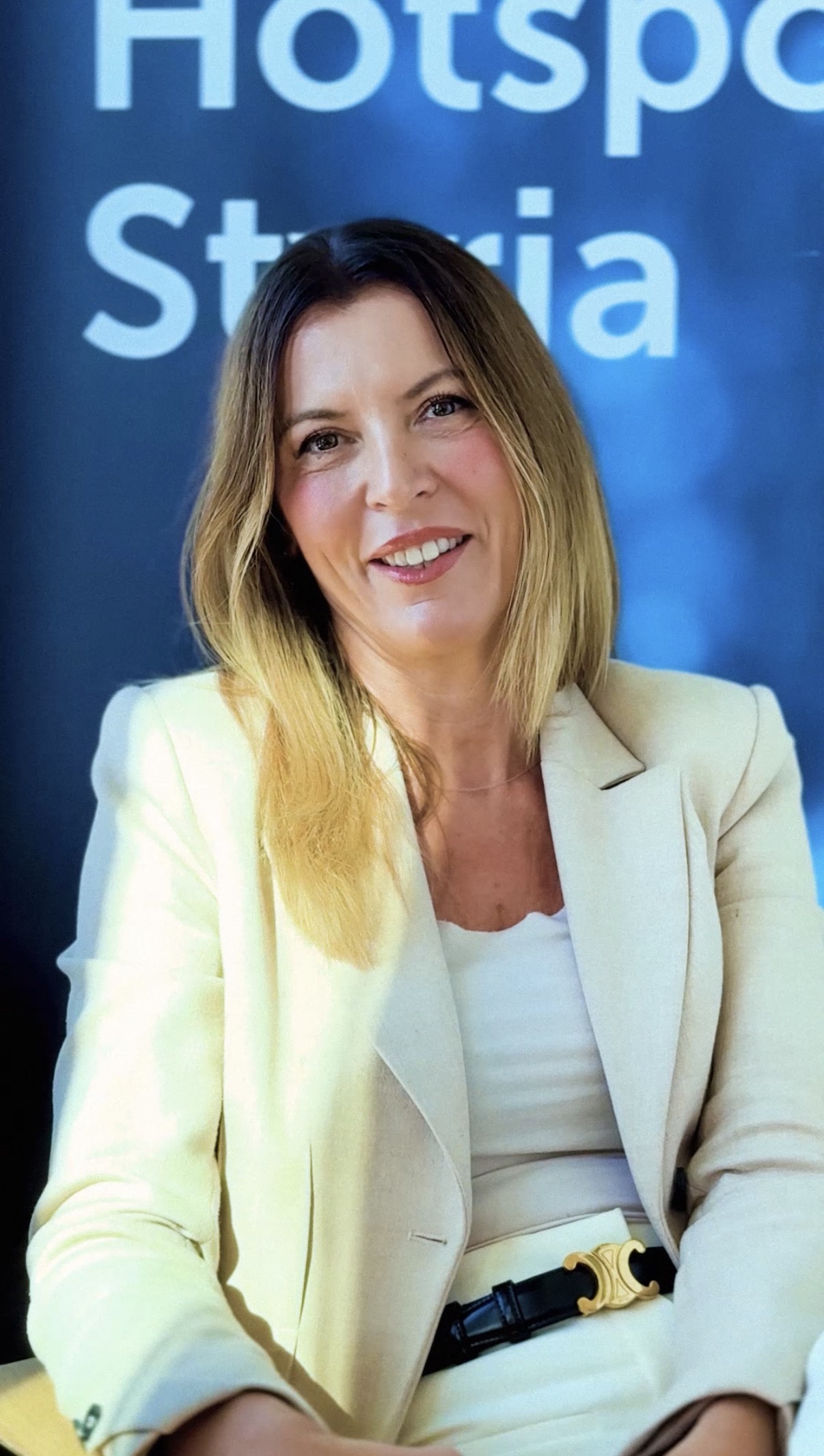 Eine Frau mit glattem, schulterlangem blondem Haar in einem cremefarbenen Blazer und weißem Oberteil lächelt, während sie vor einem blauen Hintergrund sitzt.