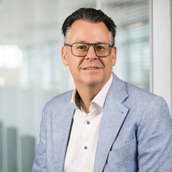 Ein Mann mittleren Alters, der eine Brille, einen hellblauen Blazer und ein weißes Hemd trägt, sitzt in einem hellen, modernen Büro mit Glaswänden und lächelt.