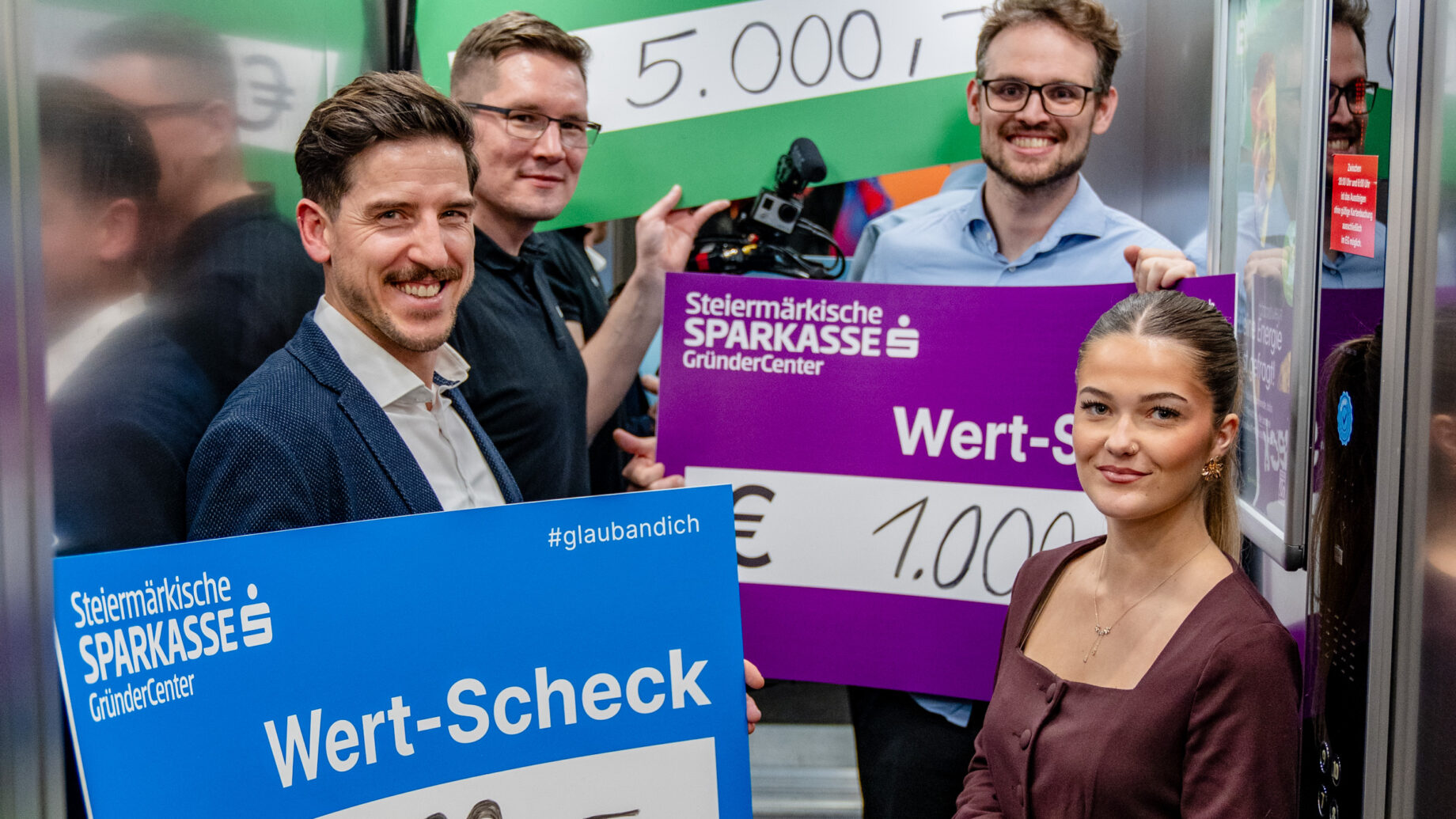 Fünf lächelnde Menschen stehen in einem Aufzug und halten große Schecks mit dem Logo der Steiermärkischen Sparkasse in der Hand, die die Ortskernbelebung Steiermark mit Beträgen von 2.000 €, 5.000 €, 1.000 € und 500 € unterstützen.