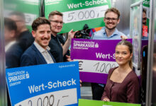 Fünf lächelnde Menschen stehen in einem Aufzug und halten große Schecks mit dem Logo der Steiermärkischen Sparkasse in der Hand, die die Ortskernbelebung Steiermark mit Beträgen von 2.000 €, 5.000 €, 1.000 € und 500 € unterstützen.