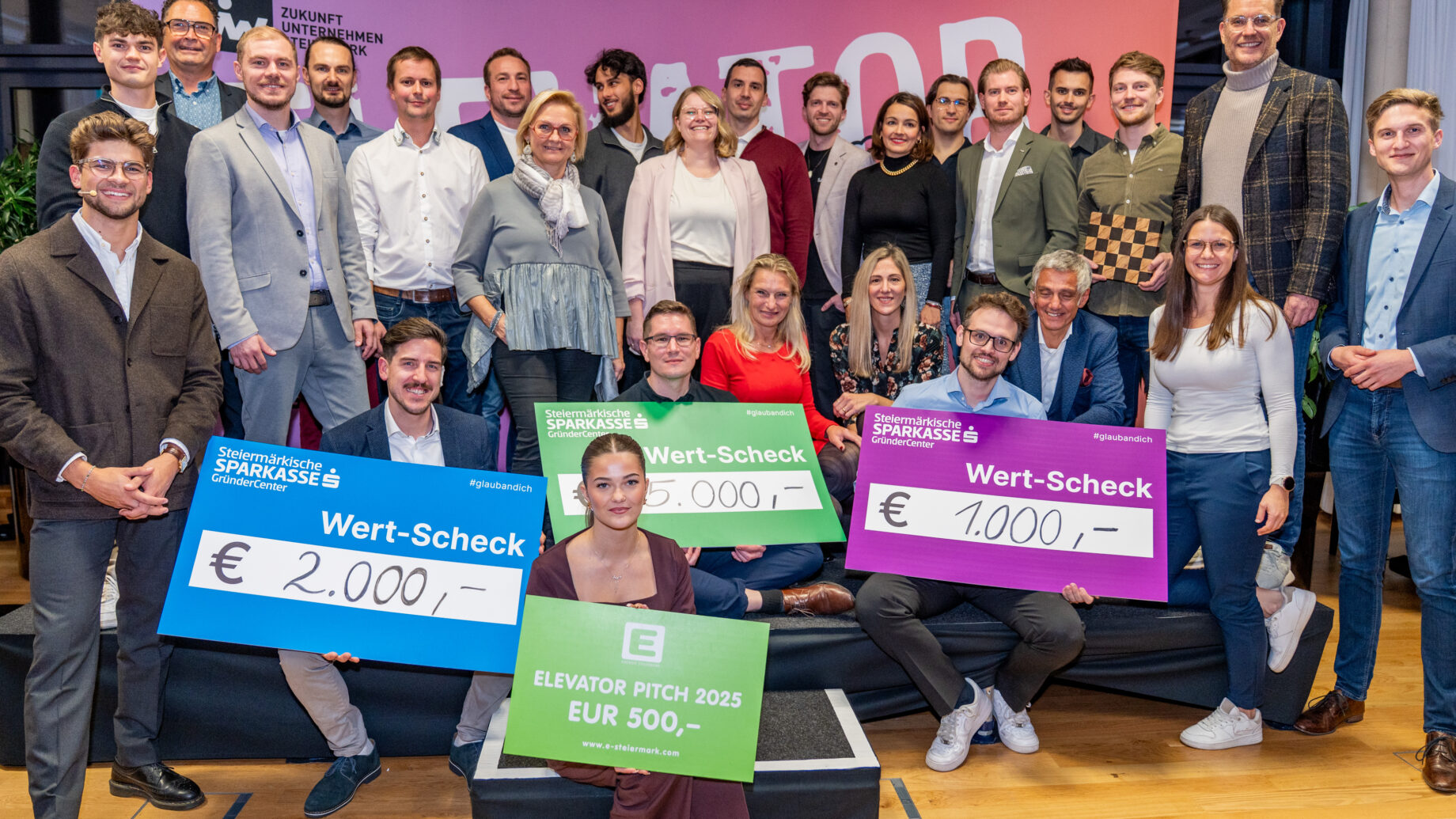 Eine Gruppe von Personen posiert in einem Innenraum bei einer Veranstaltung der Ortskernbelebung Steiermark. Drei von ihnen halten große Preisschecks über 2.000 €, 1.500 € und 1.000 € in der Hand. Vorne sitzt eine Frau mit einem 500-Euro-Scheck. Alle lächeln.