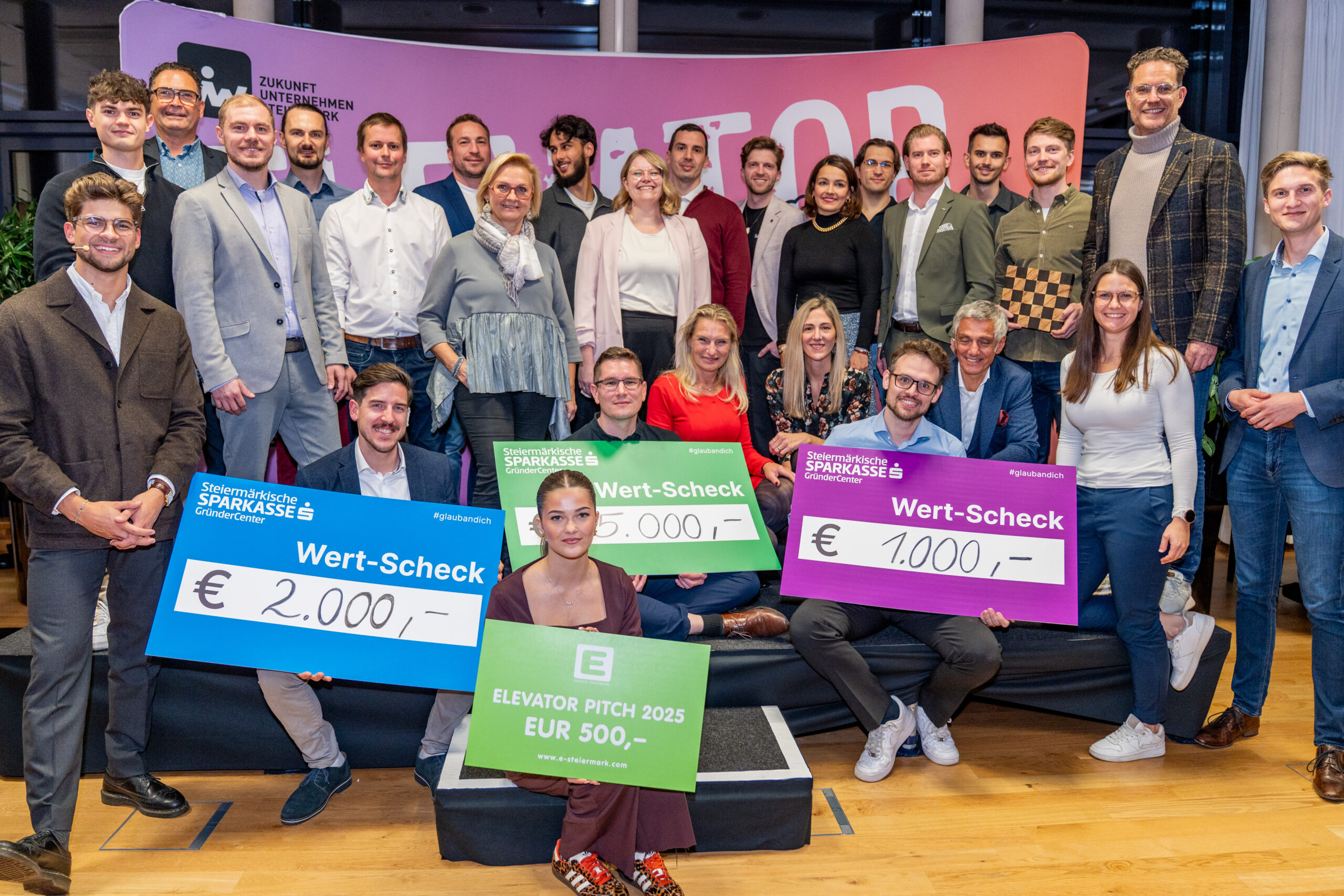 Eine Gruppe von Personen posiert in einem Innenraum bei einer Veranstaltung der Ortskernbelebung Steiermark. Drei von ihnen halten große Preisschecks über 2.000 €, 1.500 € und 1.000 € in der Hand. Vorne sitzt eine Frau mit einem 500-Euro-Scheck. Alle lächeln.