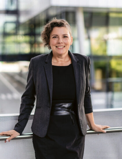 Eine Frau in schwarzem Kleid und Blazer steht im Freien, lächelt und lehnt sich an ein Metallgeländer, im Hintergrund sind moderne Glasgebäude zu sehen.