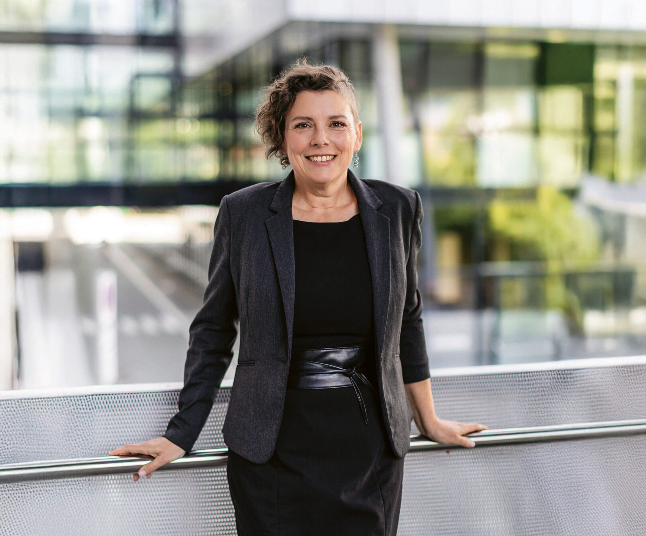 Eine Frau in schwarzem Kleid und Blazer steht im Freien, lächelt und lehnt sich an ein Metallgeländer, im Hintergrund sind moderne Glasgebäude zu sehen.