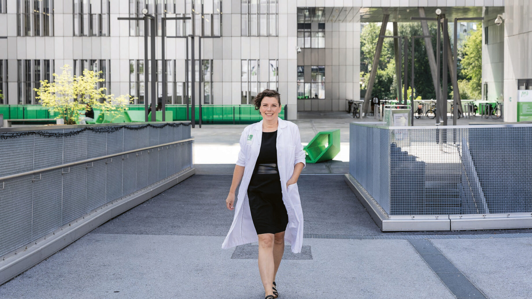 Eine Frau in schwarzem Kleid und weißem Laborkittel geht bei hellem Tageslicht selbstbewusst vor einem modernen Gebäude mit großen Fenstern und geometrischen Strukturen entlang.