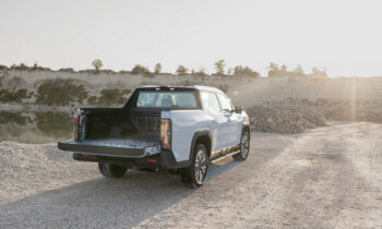 Ein weißer Pickup mit heruntergeklappter Heckklappe steht bei Sonnenuntergang auf einer Schotterstraße in der Nähe einer felsigen Landschaft.