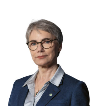 Eine ältere Frau mit kurzen grauen Haaren und Brille, die einen blauen Blazer und eine helle Bluse trägt, posiert vor einem weißen Hintergrund.