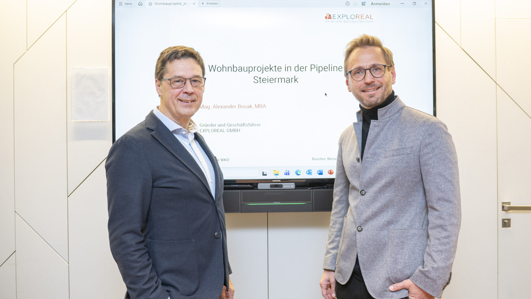 Zwei Männer in Geschäftskleidung stehen lächelnd vor einem Bildschirm, auf dem eine Präsentation mit dem Titel "Wohnbauprojekte in der Pipeline Steiermark" zu sehen ist, die das steirische Landeswappen zeigt. Die Umgebung scheint ein modernes Büro oder ein Konferenzraum zu sein.