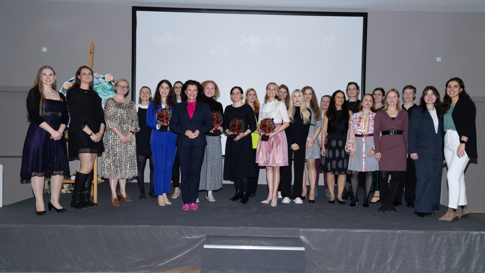 Eine Gruppe von Frauen steht auf einer Bühne, einige halten Auszeichnungen in der Hand und posieren für ein Gruppenfoto bei einer offiziellen Veranstaltung. Im Hintergrund ist eine Projektionsfläche mit Logos und Text zu sehen.