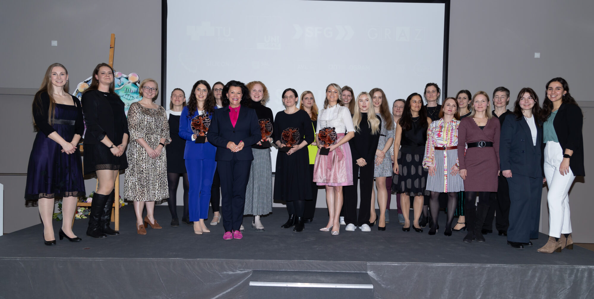 Eine Gruppe von Frauen steht auf einer Bühne, einige halten Auszeichnungen in der Hand und posieren für ein Gruppenfoto bei einer offiziellen Veranstaltung. Im Hintergrund ist eine Projektionsfläche mit Logos und Text zu sehen.