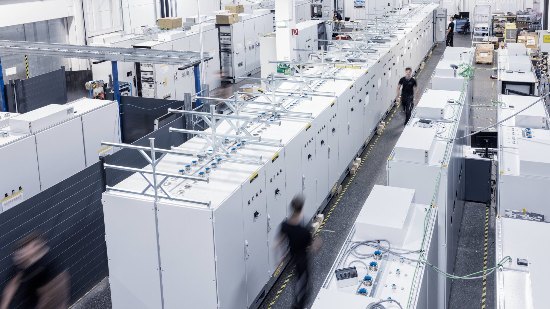 Eine große Industrieanlage mit Reihen weißer Schaltschränke und unscharfen Arbeitern, die die Gänge entlanglaufen und die Bewegung widerspiegeln. Der hell erleuchtete Bereich weist hohe Decken und Geräte auf, die den innovativen Geist der Ortskernbelebung Steiermark widerspiegeln.