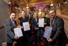 Sechs formell gekleidete Personen posieren auf einer großen Treppe in einem Innenraum und halten stolz Urkunden und Auszeichnungen in die Höhe, darunter das steirische Landeswappen der Merkur Versicherung. Im Hintergrund sind kunstvolle Lichter, ein roter Teppich und dekorative Architektur zu sehen.