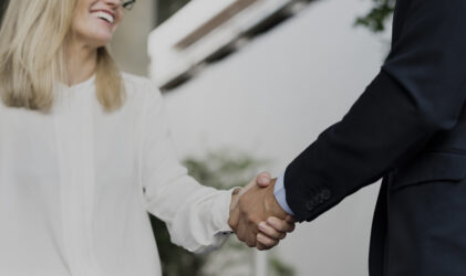 Eine lächelnde Frau in einer weißen Bluse schüttelt einer Person in einem dunklen Anzug die Hand, was auf ein berufliches oder geschäftliches Treffen hindeutet.