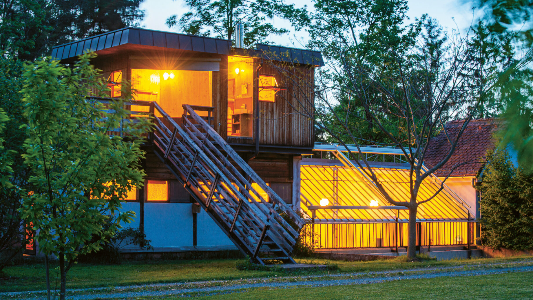 Ein modernes zweistöckiges Haus mit großen Fenstern und warmer Innenbeleuchtung, umgeben von Bäumen und Grün in der Abenddämmerung.