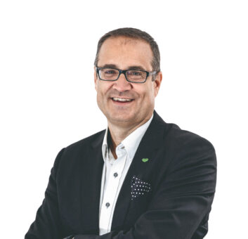 Mann mit Brille, dunklem Blazer und weißem Hemd, lächelnd mit verschränkten Armen vor einem weißen Hintergrund.