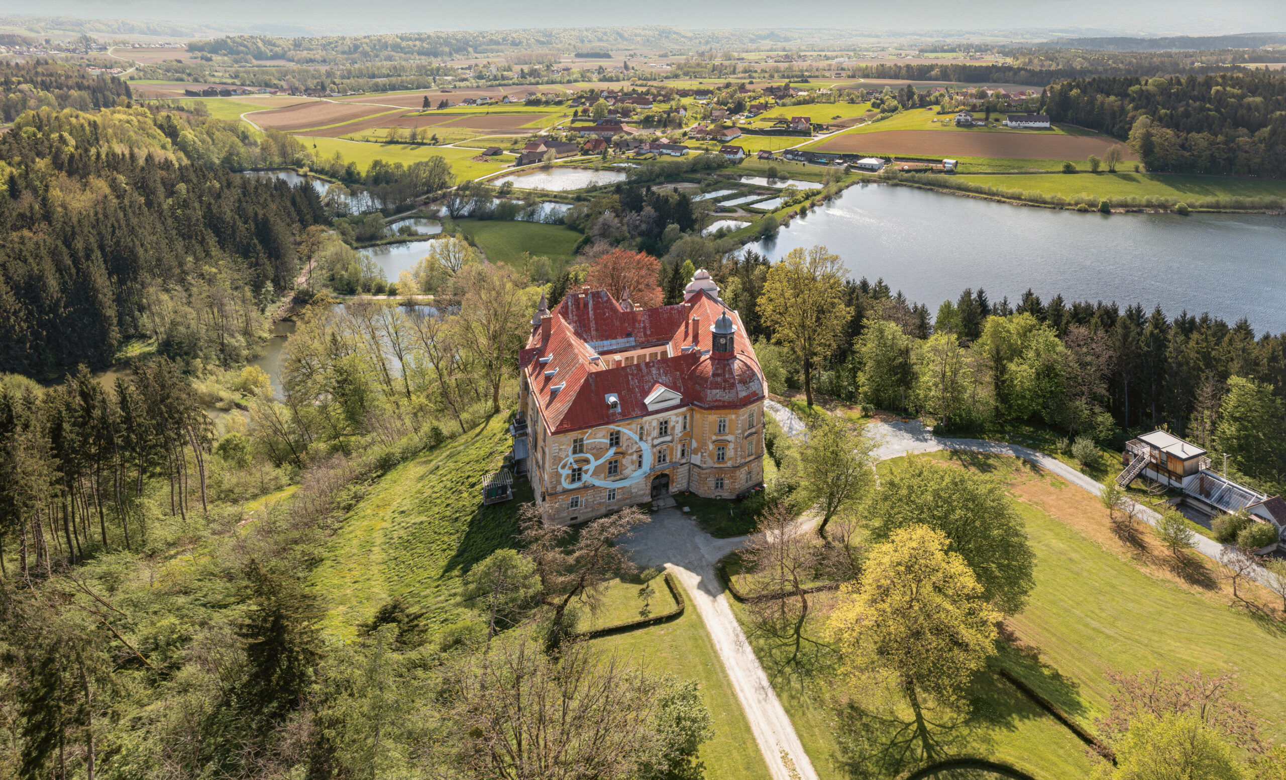Luftaufnahme eines großen historischen Herrenhauses mit rotem Dach, umgeben von Bäumen, Feldern, einer kurvenreichen Straße und einem See unter einem klaren blauen Himmel.
